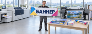 Цифровая типография: работник держит баннер, на столе плакаты, листовки и рулонный баннер, оборудование Xerox и Ricoh на фоне